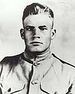 Head and shoulders of a square-jawed young man with short, light, hair, which stands straight up in the front. He is wearing a military jacket with breast pockets and a row of buttons down the center.