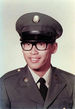 Head and shoulders of a young man wearing a peaked cap, black thick-rimmed glasses, and a military jacket with a round pin on each lapel over a shirt and tie.