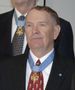 Head and shoulders of an older man with thinning hair wearing a dark suit coat, white shirt, and red tie. A star-shaped medal hangs from a light blue ribbon around his neck and on his lapel is a small round blue pin.