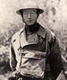 Head and torso of a man wearing a metal helmet with wide brim, wire frame glasses, a military coat, and a satchel loosely strapped to his chest.