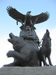 A life-sized bronze statue of an Aboriginal and eagle above him; there is  a bear to his right and a wolf to his left, they are all looking upwards towards a blue and white sky