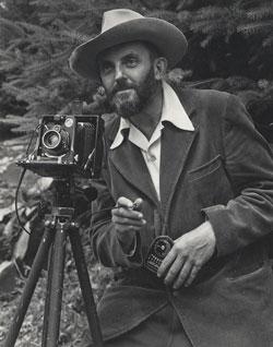 A photo of a bearded Ansel Adams with a camera on a tripod and a light meter in his hand.  Adams is wearing a dark jacket and a white shirt, and the open shirt collar is spread over the lapel of his jacket.  He is holding a cable release for the camera, and there is a rocky hillside behind him.  The photo was taken by J. Malcolm Greany, probably in 1947.