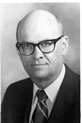 Black and white photo of a bald man wearing glasses and a suit with a striped tie