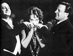 A woman, staring directly at camera, stands between two formally dressed, smiling men who gaze admiringly at her. Each is raising a hand, which she holds and kisses.