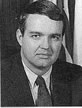 Black and white photo of a man in a suit and combed-over hair with the US flag behind him