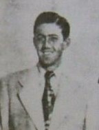 A black-haired man in a white suit and a black tie smiles as he poses for the picture