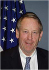 Smiling man with thinning hair wearing a suit and a blue tie with the US flag behind him