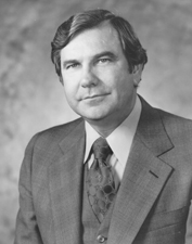 A man in his late forties with wavy black hair, facing left, wearing a suit and tie