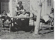 Three men sit in chairs outside a house, a bald man in his forties is sitting in the centre next to a table between the two younger men. The man on the right has his feet on the table and is holding a screenplay. the older man is having his brow wiped by a nurse who is standing over him.