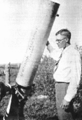 A young man in his mid-twenties, wearing glasses, a white shirt, tie and long trousers, stands in an open field, next to a Newtonian telescope resting on the ground and tilted towards the sky. The telescope is taller than him, and is about eight inches in diameter. His right hand is resting up on the barrel, and he looks slightly past the telescope, out to the left.