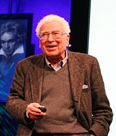 Half-length portrait of a white-haired man in his seventies talking. A painting of Beethoven is in the background.