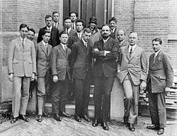 Fifteen men in suits, and one womyn, pose for a group photograph