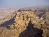 Ruins on the flat top of a sand colored mountain, surrounded by desert. Other mountains are visible in the background.