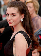 A bust shot of a young woman standing in a side view, her head turned to look to the camera. Her long hair is pulled back from her face and cascades down her back. She wears a black sleeveless dress with a gold trim around the back and under her arm. She wears jeweled silver floral shaped earrings and smiles softly. There is a crowd of people, slightly out of focus, in the background.