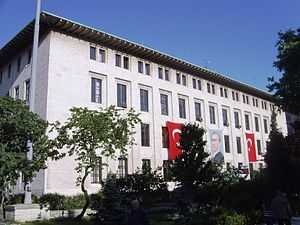 A four-story, white flat-roofed building with two Turkish flags and a portrait on the exterior