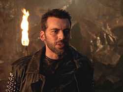 Upper body shot of bearded man in black shirt and black leather jacket, a fiery torch on the cave wall behind him