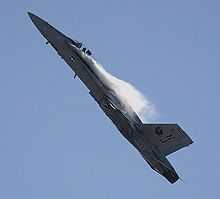 Jet fighter aircraft is seen against blue sky executing a pull-up, making it nearly vertical with contrail formed aft of the canopy.