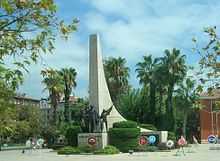 A tall sweeping stone triangle projects skyward behind the statues of a man and two children in bronze on a smaller podium. Around the base are placed several wreaths with logos. Palm trees surround the scene.