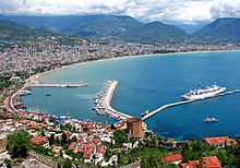 A colorful city with red roofs rising out from a curving harbor with blue water and cruise ship docked by a long pier.