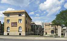 From left to right, a yellow brick building with a flat roof; a steel, concrete and glass connector, and another brick building with a large tree and lawn in front, seen from across a city street.