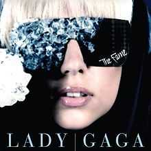 The face of a blond woman. She is wearing black glasses. The right side of the glasses is covered by blue crystals. On the bottom of the left side of the glasses, the word "The Fame" is inscribed in white.