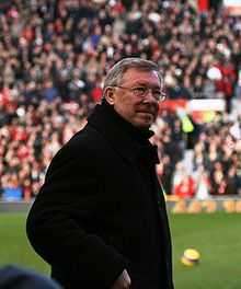 The torso and head of a grey-haired white man. He is wearing spectacles and a black coat.