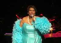 A woman stands on stage and sings into a microphone. She wears a blue dress and has a blue feather boa around her arms.