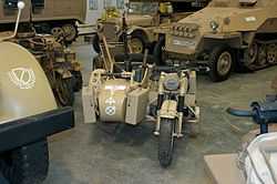 Located in a museum among other vehcicles, a front view of a desert camouflage BMW R75 motorcycle and sidecar