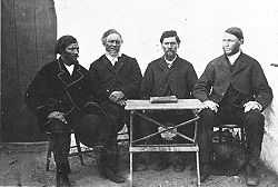 A black-and-white photography from 1872. Four middle-aged men sit behind a small folding table in front of a cottage. A book is prominently placed in the middle of the table.