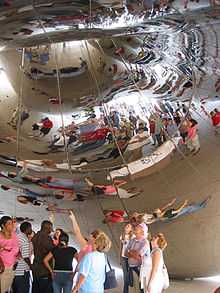 The reflections of visitors are warped in the mirror-like surface of an object above them; the surface of the object is covered in weld lines.