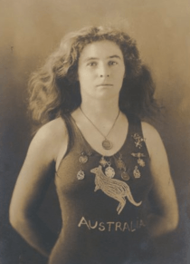 A sepia-toned photograph taken from the waist up, showing a white woman of medium build with shoulder-length wavy hair, wearing a one-piece swimming costume with the word "AUSTRALIA" on the front and a depiction of a kangaroo. She has nine medals pinned to costume around the upper chest area, and is wearing another medal around her neck.