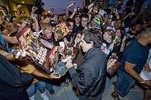 A crowd of people is all looking towards a man at the center who is signing a hat. The crowd is attempting to hand him posters to sign and others are taking pictures using cameras and cell phones.