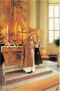 "A partially bald Bo Giertz with glasses is shown wearing a gold cope over his red stole and white cassock; in his right arm he holds a white and gold mitre; his left hand clasps his shepherd's crozier; he is standing on a raised step before an ornately decorated altar with candles and crucifix and is facing the congregation, who are not seen in the picture."