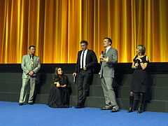 Scott Franklin, Mila Kunis, Vincent Cassel, Darren Aronofsky, and Sandra Hebron stand on a stage with a golden curtain backdrop wearing formal attire and discussing Black Swan