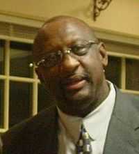 A black person, wearing a gray suit, a tie and glasses, is looking to the front.