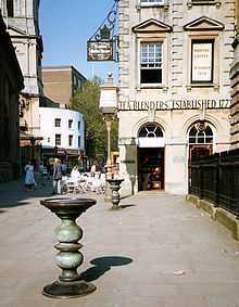 Two ornate metal pillars with large dishes on top in a paved street, with an eighteenth-century stone building behind, upon which can be seen the words "Tea Blenders Estabklishec 177-". People sitting at cafe-style tables outside. On the right iron railings.