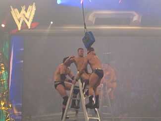 Three Caucasian professional wrestlers fight on top of a ladder in the ring. The wrestler in the centre in reddish-maroon trunks is about to elbow the wrestler on the right. A blue briefcase hangs in the air barely above the three wrestlers.