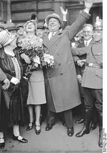 A man wearing a hat and a long coat and pants, he is surrounded by people.