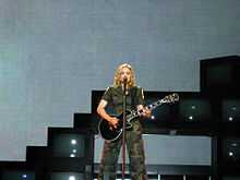 A blond woman, wearing olive green clothes stands in front of a gray backdrop and sings to a microphone while holding a black electric guitar in her hands. In the background a structure made out of television sets can be seen