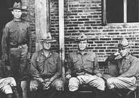 Four men in military uniforms wearing hats. Three are seated on a bench and one is standing behind the others.