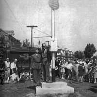 A man in a suit standing on a stage next to a large pole. There is a Marine in the background in his dress uniform and behind him a crowd of people are watching the man on the stage.