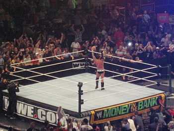 A Caucasian professional wrestler  stands alone in the middle of the ring, hoisting a golden championship belt. The ring canvas at the side of the ring reads "WWE.com" and "Money in the Bank". The crowd at ringside appears to be celebrating with the wrestler.