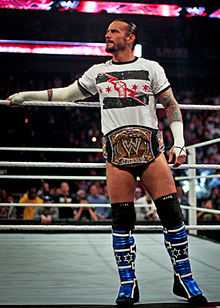 A black-haired Caucasian man in a white T-shirt stands on the ring apron, a golden belt with the WWE insignia around his waist.