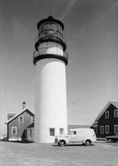 High-resolution black and white photo of the lighthouse and keeper's house.  Parked in front of the lighthouse is a 1950s van, with a military emblem and the words "United States Coast Guard" stenciled on the driver's door, and on the side of the van is a military advertisement that reads "The U.S. Coast Guard NEEDS MEN! Join Now! No waiting".