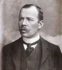  Head and shoulders portrait of a man with receding hair, heavy moustache, looking left from the image. He wears a high white collar, black necktie, dark waistcoat and jacket.