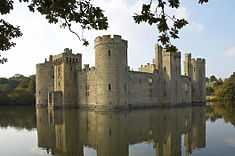 Photo of Bodiam Castle across the water, framed by oak leaves. The alternation of large round towers at the corners and square towers central to each face can be seen. Daylight visible through upper windows indicates that the roofs and inner structures are ruined.