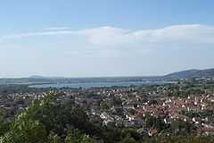 Roofs of multiple buildings separated by trees and vegetation. In the distance is a lake and hills.