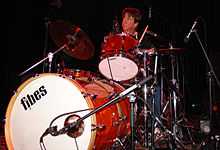 Chris Layton playing a drum kit. He has dark hair and is looking upwards.