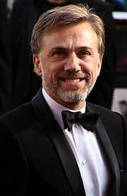 A dark blond haired man in a black tux and bowtie and white shirt faces forward while smiling.