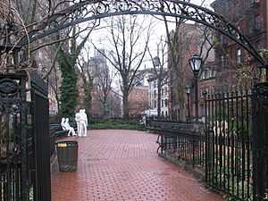 A color photograph of Christopher Park in winter, showing the wrought iron entrance arch in the foreground and the brick pavement surrounded by five and six story brick buildings; in the center background are four white statue figures: two males standing, one with his hand on the other's shoulder, and two females seated on a park bench, one woman with her hand touching the other's thigh. All are dressed in jeans and loose clothing